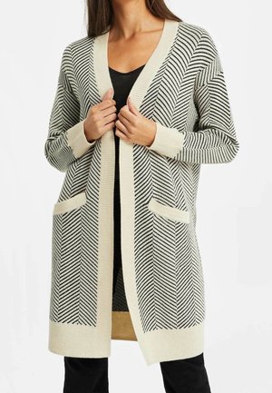 Femme portant un long cardigan en maille crème et noire à motif chevrons avec des poches avant, par-dessus un haut noir et un pantalon noir, tenant les bords du cardigan.