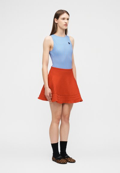 Femme debout, portant un haut sans manches à rayures bleues, une jupe évasée rouge, des chaussettes noires et des chaussures à imprimé léopard sur un fond blanc uni.