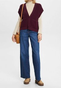 Burgundská pletená vesta se dvěma předními kapsami přes béžový rolák, kombinovaná s modrými džínami s širokými nohavicemi a kotníkovými botami. Zahrnutý je také hnědý kabelka přes rameno.