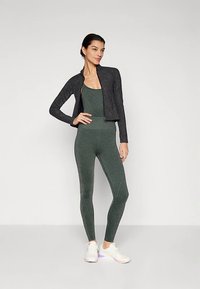 Mörkgrå kort jacka över en grön, glansig sport-bodysuit med matchande tights. Modellen bär vita sneakers med rosa detaljer.
