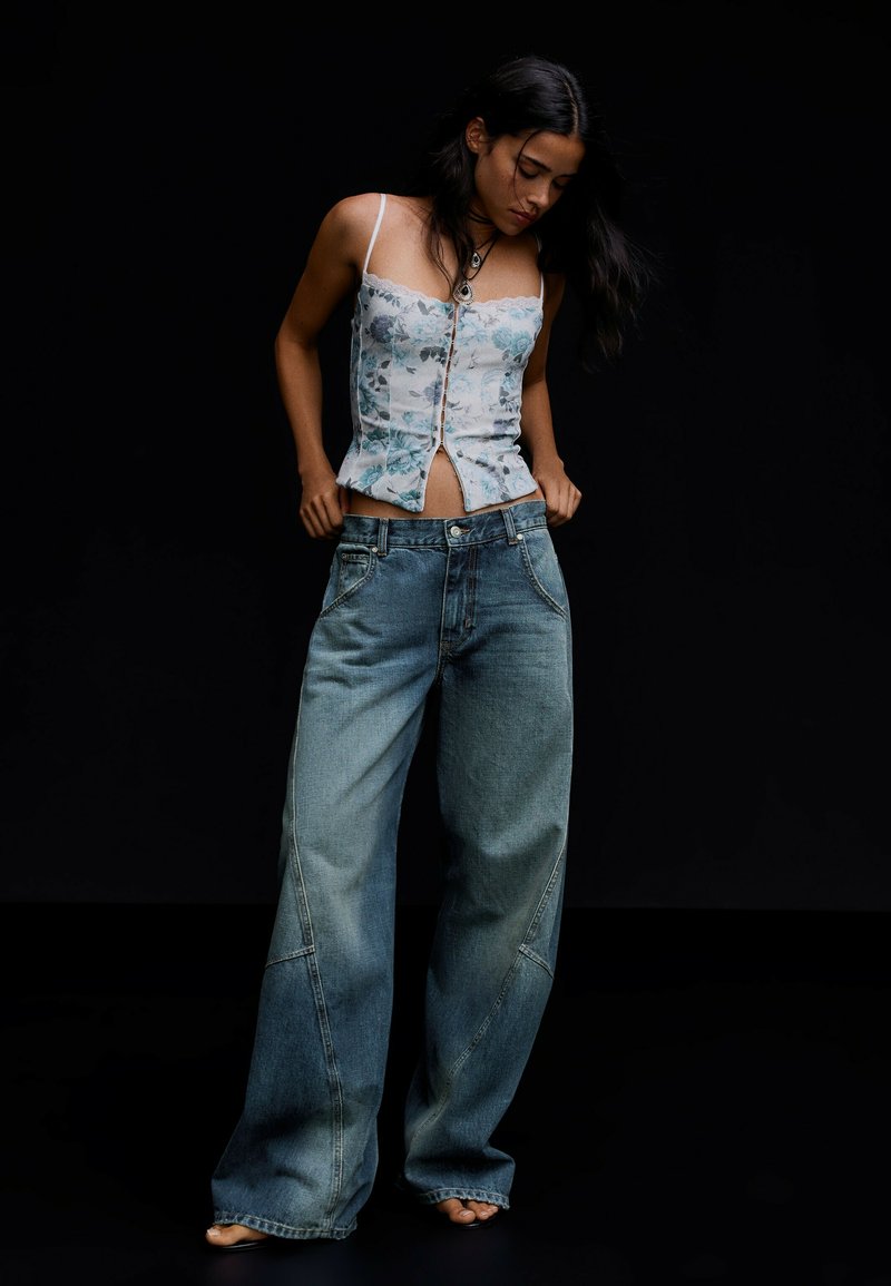 Femme aux longs cheveux foncés portant un corset à fleurs et un jean bleu oversize, posant pieds nus sur fond noir.