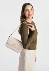 Beżowa torebka syntetyczna z złotym logo, prostokątna kształt i regulowany pasek, noszona na ramieniu, w zestawieniu z ciemnozieloną bluzką.