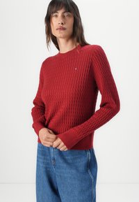 Suéter rojo de punto con un patrón texturizado, cuello redondo y puños acanalados, combinado con pantalones de mezclilla azul. Incluye un pequeño detalle de logo.