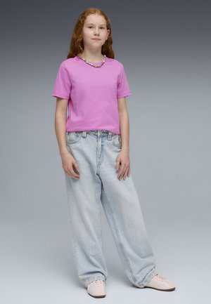 Joven con cabello largo y rojo lleva puesta una camiseta rosa, jeans de pierna ancha azul claro, zapatillas rosas y un collar colorido de cuentas.