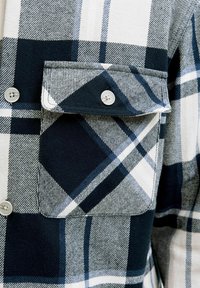 Camisa de cuadros en blanco y negro con un bolsillo en el pecho, cierres de botón y una textura de tela suave con patrones variados.