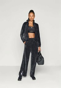 Μαύρο φούτερ παντελόνι με ζακέτα με φερμουάρ και φαρδιά παντελόνια. Το σύνολο έχει απαλή υφή και από κάτω φέρει ένα εφαρμοστό μαύρο σουτιέν γυμναστικής.