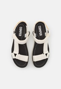 Paire de sandales noires avec des brides blanches réglables et la marque "Camper" sur les semelles intérieures, vues de dessus.