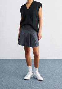 Svart stickad ärmlös topp med V-ringning, ihop med en grå plisserad kjol och vita sneakers, mot en neutral bakgrund.