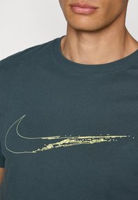 Temno turkizna bombažna majica s velike rumene Nike swoosh grafike. Okrogli ovratnik, kratke rokave, gladka tekstura in sproščen kroj.