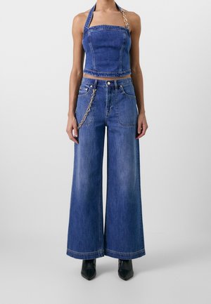 Kobieta w szerokich niebieskich dżinsach z denimu i dopasowanym topie na szyję z paskiem z złotych łańcuszków, czarne szpiczaste buty na szarym tle.