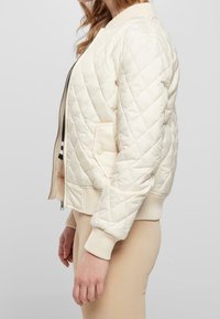 Chaqueta bomber acolchada en beige claro, con puños y dobladillo acanalados, cierre de cremallera y bolsillos laterales. Textura suave y acolchada.