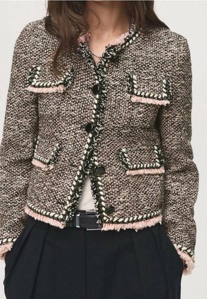 Femme portant une veste en tweed texturé avec des boutons noirs, une bordure à franges rose et noire, quatre poches avant, associée à un pantalon noir et une ceinture.