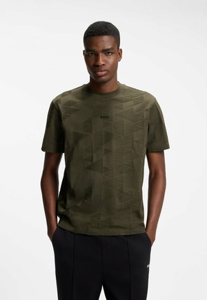Homme portant un T-shirt vert olive à motif géométrique avec le logo « BOSS » et un pantalon noir, mains dans les poches, debout devant un fond blanc.