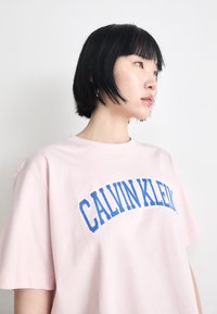 Rozā kokvilnas T-krekls ar izliekta zila "CALVIN KLEIN" emblēma uz krūtīm. Īsas piedurknes un brīvs piegriezums.