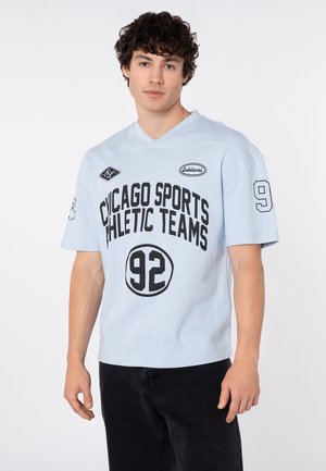 Junger Mann mit dunklen lockigen Haaren, der ein hellblaues Sportshirt mit der Aufschrift "Chicago Sports Athletic Teams 92" und schwarze Hosen trägt, stehend.