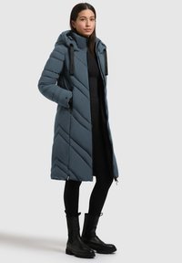 Pitkä sininen untuvatakki, jossa on soikea kuvio, vetoketjulliset taskut ja huppu. Sisältää tekstuuripintaa ja mustia metalliosia.