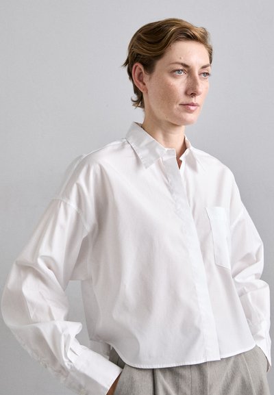 Žena s krátkymi svetlohnedými vlasmi, oblečená v voľnej bielej košeli s gombíkmi a šedých nohaviciach, pózujúca proti jednoduchému svetlosivému pozadiu.