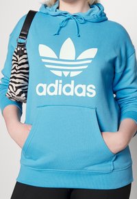Světle modrá mikina vyrobená z měkké tkaniny, s velkým bílým logem Adidas a klokanní kapsou. V kombinaci s taškou v černobíl ém zebrovém vzoru.