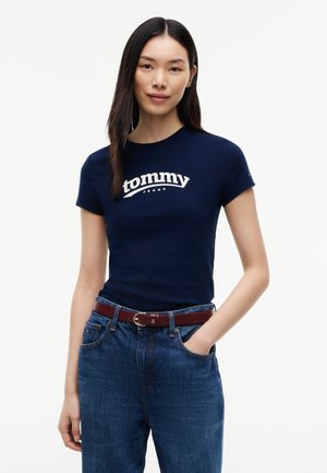 Femme aux longs cheveux foncés porte un t-shirt Tommy Jeans bleu marine, un jean bleu et une ceinture marron, debout avec une main dans la poche.