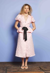 Γυναίκα με σγουρά μαλλιά, φορώντας ένα ροζ φλοράλ φόρεμα με φουσκωτά μανίκια, μαύρη ζώνη με φιόγκο, χρυσά κοσμήματα και λευκές σαγιονάρες, στέκεται σε ξύλινο δάπεδο.