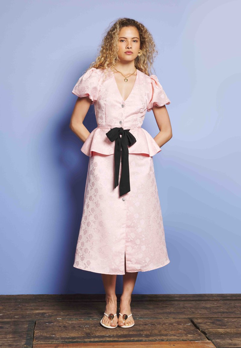 Γυναίκα με σγουρά μαλλιά, φορώντας ένα ροζ φλοράλ φόρεμα με φουσκωτά μανίκια, μαύρη ζώνη με φιόγκο, χρυσά κοσμήματα και λευκές σαγιονάρες, στέκεται σε ξύλινο δάπεδο.