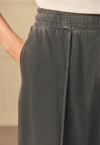Primo piano di una mano che riposa nella tasca di pantaloni grigi scuri con elastico in vita, realizzati in tessuto testurizzato e con cuciture laterali visibili.