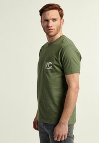 Camiseta de algodón de manga corta en color verde oliva con un bolsillo en el pecho que presenta un logo blanco "VC". Diseño clásico de cuello redondo.