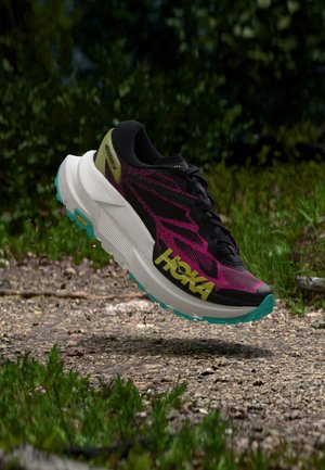 Chaussure de course noire, violette, jaune et blanche planant au-dessus d’un chemin de gravier avec des plantes vertes en arrière-plan.