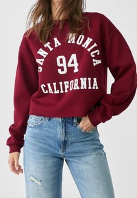 Donna che indossa una felpa bordeaux con la scritta "Santa Monica 94 California" e jeans strappati di un blu chiaro, in piedi con una mano in tasca.