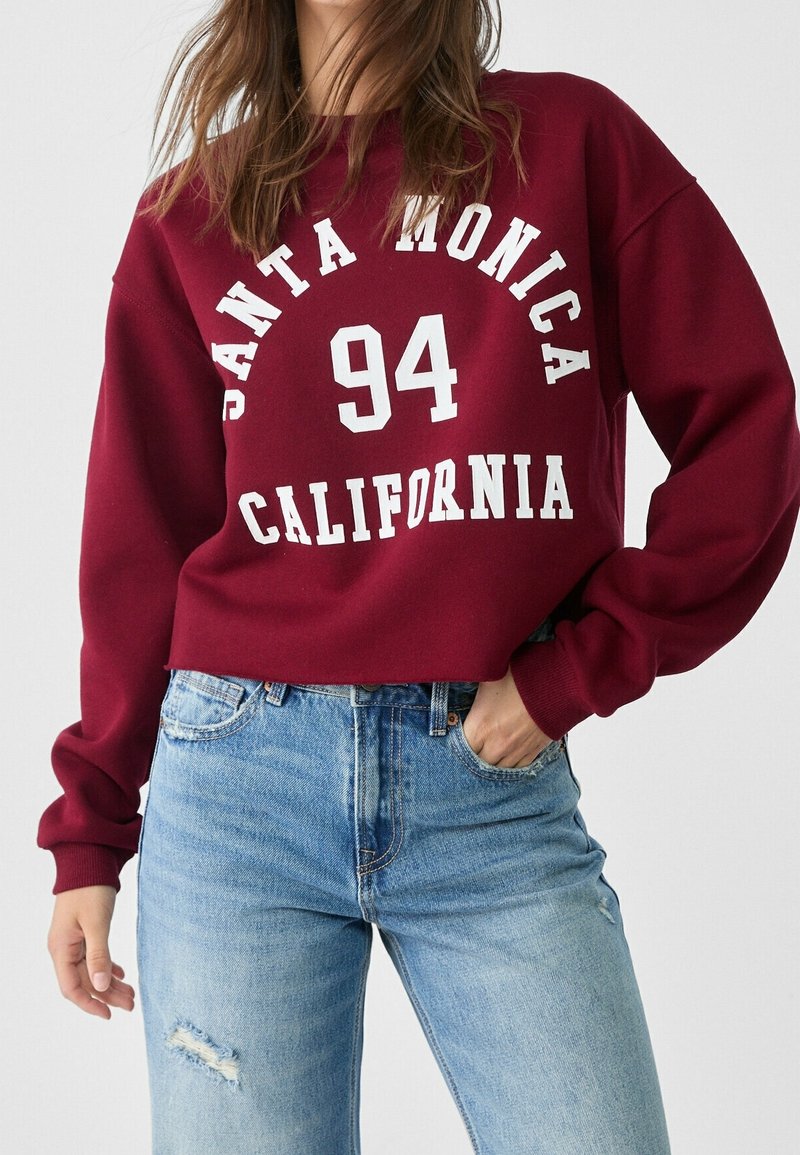 Donna che indossa una felpa bordeaux con la scritta "Santa Monica 94 California" e jeans strappati di un blu chiaro, in piedi con una mano in tasca.