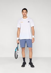 Camiseta blanca con texto naranja, logo azul y mangas cortas. Pantalones cortos azul claro, zapatillas negras, sosteniendo una raqueta de tenis. Calcetines negros.