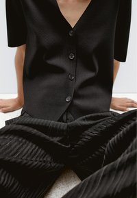 Zwart T-shirt met korte mouwen en V-hals, voorzien van een sluiting met zes knopen, gecombineerd met zwarte geplooide broek met verticale strepen en een gestructureerde stof.