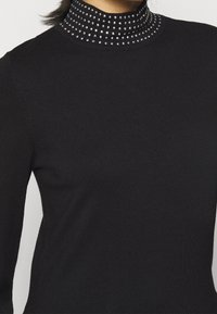 Svart högkragad tröja i strukturerad tyg med en krage prydd med metallnitar jämnt fördelade runt halslinjen.