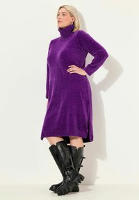 Abito a tubino viola a maglia con colletto alto e maniche lunghe; presenta delle aperture laterali. Abbinato a stivali neri in pelle al ginocchio con suole spesse e fibbie.