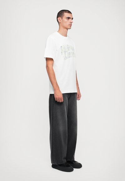 Un tricou alb din bumbac cu text grafic verde, combinat cu o pereche de pantaloni din denim capitonați, gri închis, cu croială lejeră. Modelul stă de profil, purtând pantofi negri.