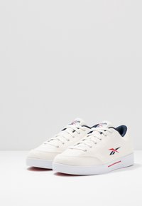 Zapatillas de lona blancas con diseño de corte bajo, detalles en marino y logo rojo. Presentan cordones redondos y una suela de goma texturizada.