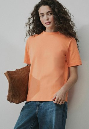 Femeie cu păr creț purtând un tricou scurt portocaliu și blugi albaștri, ținând o geantă mare de culoare maro țesută, sprijinită de un perete deschis la culoare.