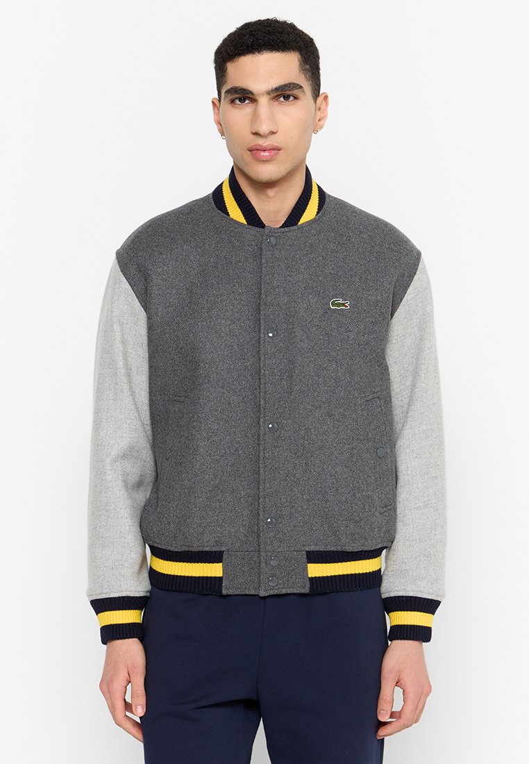 Veste de baseball grise et gris clair avec des poignets côtelés bleu marine et jaunes. Présente une fermeture à boutons et le logo Lacoste sur la poitrine.