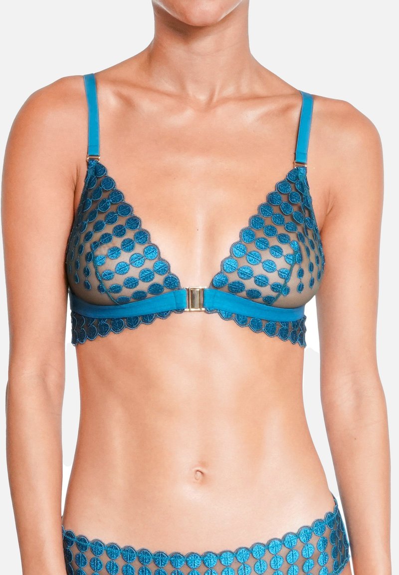 Femme portant un soutien-gorge bleu transparent à pois avec un fermoir à l'avant, montrant le ventre et une partie de la culotte assortie sur un fond blanc.