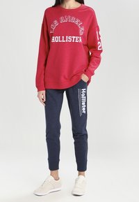 Červená mikina s bílým nápisem "LOS ANGELES HOLLISTER", doplněná tmavými námořnickými joggery s bílým nápisem "Hollister", dokončeno lehkými teniskami.