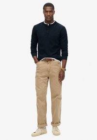 Man standing with hands in pockets, wearing navy long-sleeve henley shirt, beige cuffed pants, white sneakers, brown belt, and wristwatch.