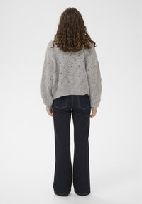 Frau mit lockigem Haar, die einen grauen Strickpullover und dunkelblaue Schlagjeans trägt, steht mit dem Rücken zum Betrachter vor einem schlichten weißen Hintergrund.