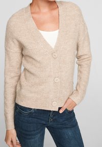 Béžový žebrovaný cardigan vyrobený z měkké pleteniny, s hlubokým V výstřihem, čtyřmi velkými knoflíky a zkráceným lemem, v kombinaci s tmavými džíny.