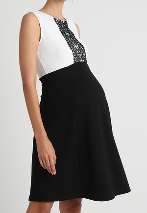 Kobieta w ciąży ubrana w czarno-białą sukienkę bez rękawów z koronkowymi detalami na piersiach na jednolitym tle.