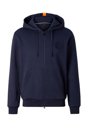 Młody mężczyzna ubrany w granatową bluzę z kapturem na suwak, dopasowane spodnie dresowe, niebieską czapkę, lustrzane okulary przeciwsłoneczne, białe skarpetki i jasnoszare sneakersy stoi nieruchomo.