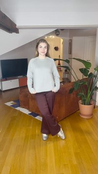 Camisola cinzento claro de manga comprida com textura canelada combinada com calças bordô de perna larga e ténis prateados; ambiente interior com plantas.
