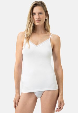 Femme aux longs cheveux blonds portant un caraco blanc et des sous-vêtements assortis, debout devant un fond clair uni.