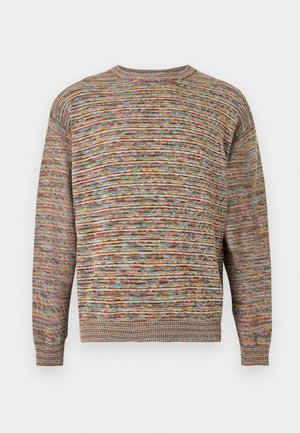 Muž s krátkymi blond vlasmi, oblečený v hnedom pruhovanom svetri, džínsových šortkách, oranžových ponožkách a bielych teniskách, stojaci s jednou rukou v vrecku.