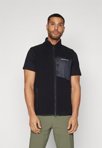 Gilet en polaire noir avec un col haut, fermeture éclair et une poche poitrine noire brillante. Porté sur une chemise noire à manches courtes avec un pantalon vert.