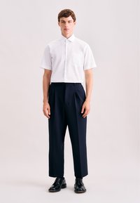 Wit overhem met korte mouwen en knoopsluiting, voorzien van een borstzak, gecombineerd met marineblauwe geplooide broek en zwarte veterschoenen. Strak en op maat gemaakt ontwerp.